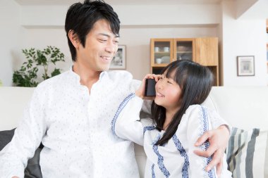 Asyalı kız babasıyla koltukta otururken akıllı telefondan konuşuyor. 