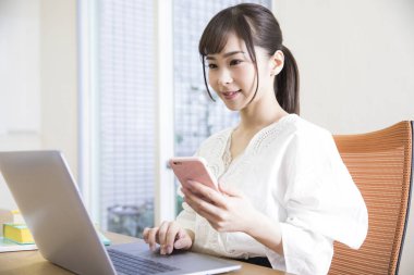 Ofiste akıllı telefon ve dizüstü bilgisayar kullanan genç Asyalı kadın..
