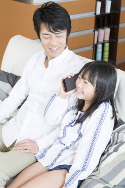 Asyalı kız babasıyla koltukta otururken akıllı telefondan konuşuyor. 