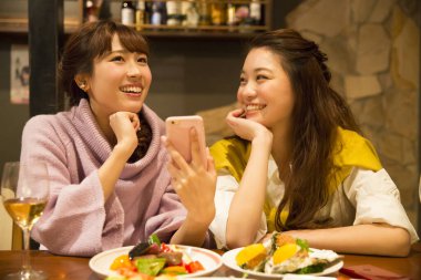 İki genç Asyalı kadın restoranda yemek yiyor, telefonda bir şeyler izliyorlar.