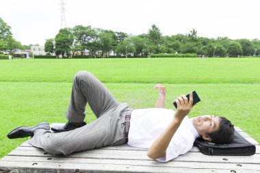 gülümseyen Asyalı iş adamı parkta bankta uzanıyor ve akıllı telefon kullanıyor.