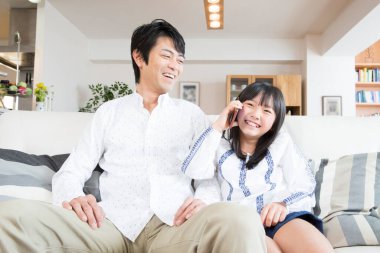 Asyalı kız babasıyla koltukta otururken akıllı telefondan konuşuyor. 