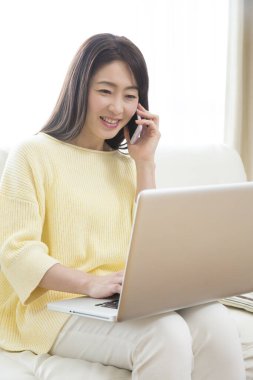 Genç Asyalı kadın dizüstü bilgisayarda çalışıyor ve evde akıllı telefonla konuşuyor. 