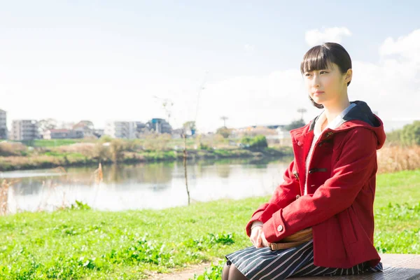 亚洲女孩穿着红色夹克坐在湖边的公园里 — 图库照片