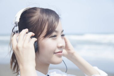 Okul üniformalı gülümseyen Asyalı öğrenci plajda kulaklıkla müzik dinliyor.