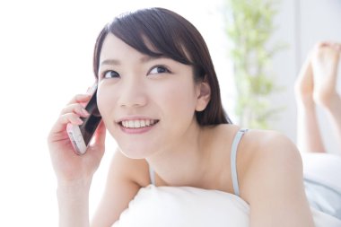 Asyalı genç bir kadın cep telefonuyla yatakta yatıyor.