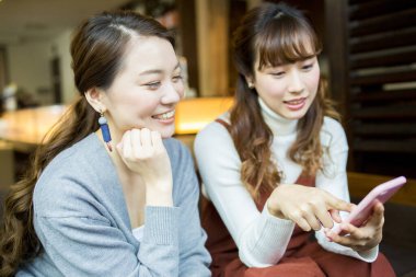 İki Asyalı kadın akıllı telefon kullanıyor ve kafede dinlenirken gülümsüyor.