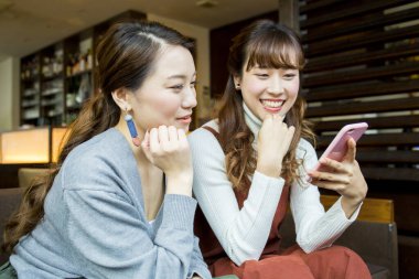 İki Asyalı kadın akıllı telefon kullanıyor ve kafede dinlenirken gülümsüyor.
