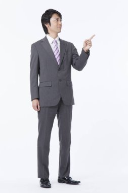 Asyalı takım elbiseli adam beyaz arka planda durup parmağıyla işaret ediyor. 