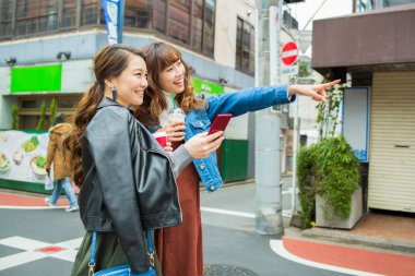 Şehirde yürüyen iki genç Asyalı kadın. Kadın arkadaşına bir şey gösteriyor. 