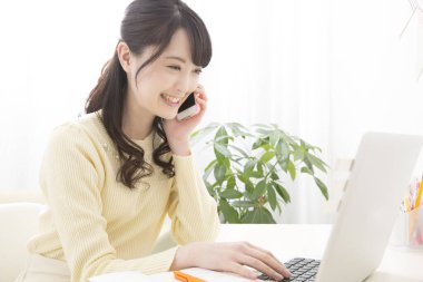 Genç Asyalı kadın dizüstü bilgisayar kullanıyor ve evde akıllı telefonla konuşuyor.