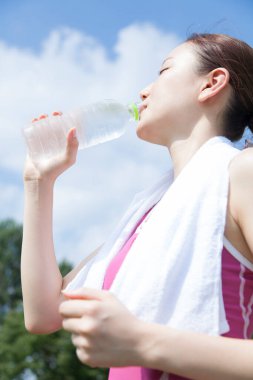 Genç Asyalı kadın parkta antrenman yaptıktan sonra şişeden içiyor. 