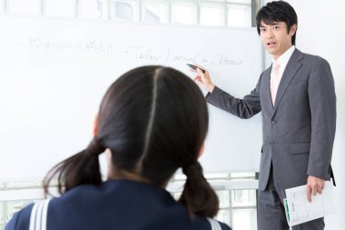 Asyalı erkek öğretmen öğrencilere tahtanın yanında duran materyalleri açıklıyor.