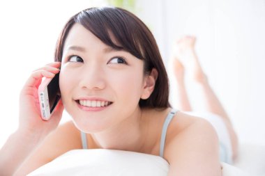 Genç Asyalı kadın akıllı telefonuyla yatakta yatıyor. 