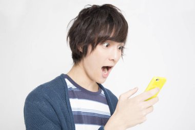 Genç Asyalı adam beyaz arka planda izole edilmiş cep telefonunu kullanırken şok oldu.