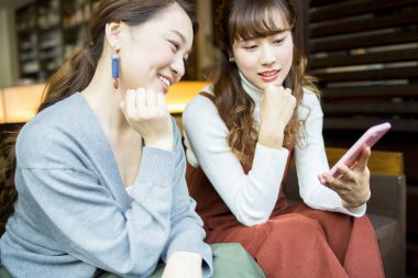 İki Asyalı kadın akıllı telefon kullanıyor ve kafede dinlenirken gülümsüyor.