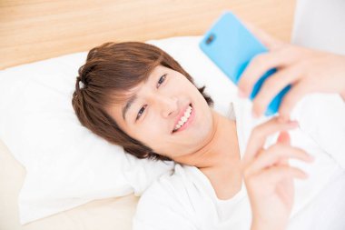 Genç Asyalı adam akıllı telefonuyla yatakta yatıyor.