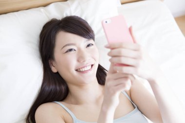 Genç Asyalı kadın gülümseyen ve yatakta uzanan akıllı telefon kullanıyor.