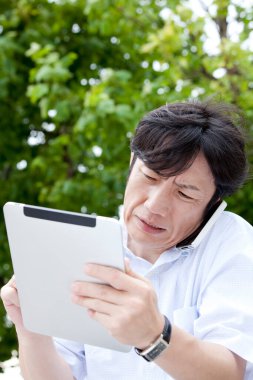 Asyalı adam tablet kullanıyor ve parkta akıllı telefondan konuşuyor. 