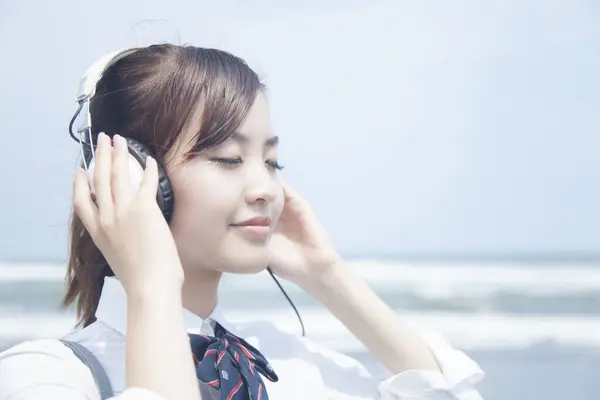 身穿校服 面带微笑的亚洲学生 带着耳机在沙滩上听音乐 — 图库照片