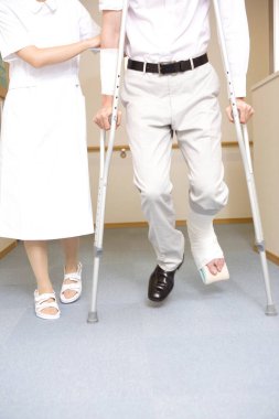 Yaralı adam, hemşirenin yanında koltuk değneği kullanarak yürüyor.