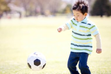 Küçük çocuk yeşil alanda futbol oynuyor.