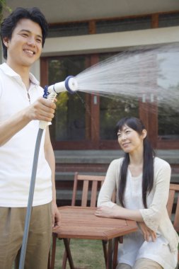 Kadın izlerken erkek bahçeyi suluyor.