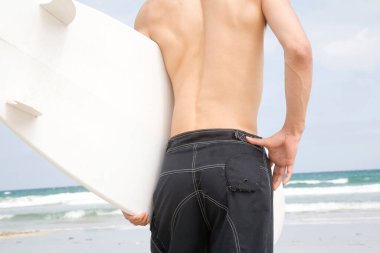 Sahilde sörf tahtası olan Asyalı genç bir adamın arka görüntüsü.