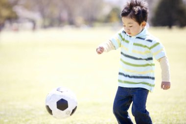 Küçük çocuk yeşil alanda futbol oynuyor.