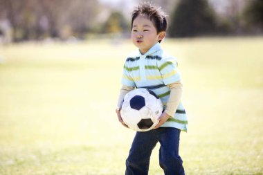 Küçük çocuk yeşil alanda futbol oynuyor.