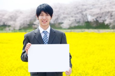 Yakışıklı Asyalı genç iş adamının portresi boş beyaz levhalı çiçek açan kolza tarlasının yanında duruyor.