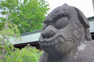 Yıpranmış taş aslan heykeli yeşil yapraklı ağaçların yakınındaki girişi koruyor.