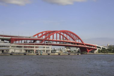 Büyük kırmızı kemer köprüsü mavi gökyüzünün altında geniş bir nehri kaplıyor.