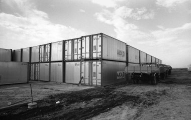 Bulutlu gökyüzünün altındaki inşaat sahasına metal nakliye konteynerleri yığılıyor.