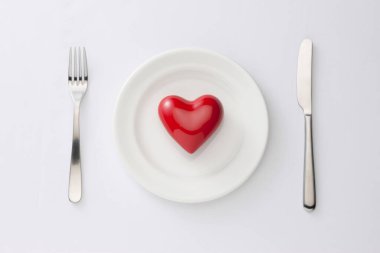 Kırmızı kalp beyaz tabakta, çatal ve bıçak beyaz arkaplanda.