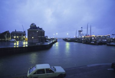 Akşamki liman sahnesinde sakin suyun yanında park etmiş bir araba var.