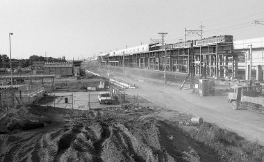 Ağır makineler ve tamamlanmamış yüksek yapılar içeren tozlu bir şantiye.