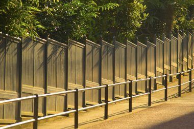 Güneş ışığı yürüme yolunun yanında metal çitler boyunca gölgeler bırakıyor.