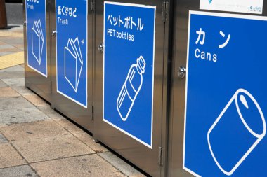 Açık havada Japon etiketleri ve beyaz simgeleri gösteren mavi geri dönüşüm kutuları.