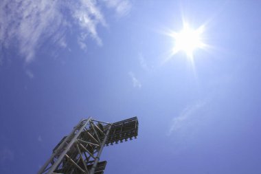 Açık mavi gökyüzüne karşı stadyum ışıklarının üzerinde parlayan parlak güneş.