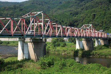 Nehir boyunca uzanan yeşil tepelerle çevrili metal kiriş köprüsü.