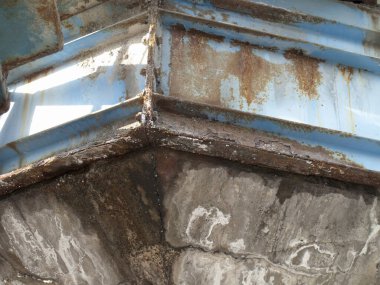 Yıpranmış beton yapı, dışarıda paslı mavi metal kirişlerle buluşuyor.
