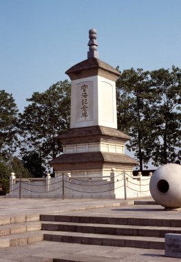 Mavi gökyüzünün altındaki ağaçların arasında duran tarihi taş heykel.