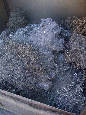 Büyük metal konteynırın içinde yanmış dalları kaplayan gri küller.