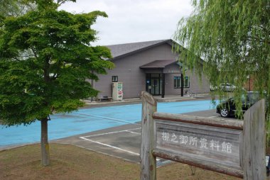 Mavi otoparkın yanındaki ahşap bina yeşil ağaçlarla çevrili.