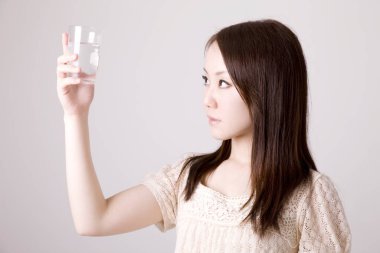 Açık arkaplanda bir bardak suyla güzel bir Asyalı kadının yakın plan fotoğrafı.