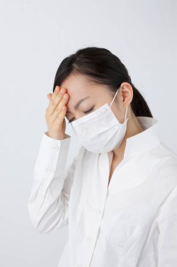 Maske takan, baş ağrısı çeken genç Asyalı kadın beyaz arka planda izole edilmiş. 