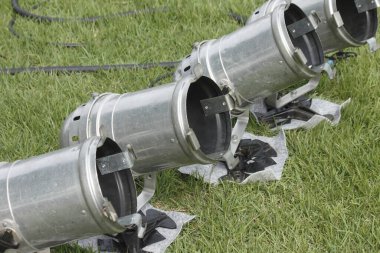 Dışarıdaki yeşil çimlerin üzerinde köşegen şeklinde duran dört metal sahne ışığı.