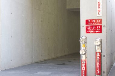 Beton koridorda kırmızı acil durum işaretleri ve yangın musluğu var.