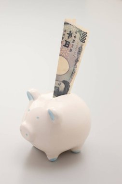 Beyaz domuz kumbarası Japon yen banknotunu dik tutuyor.